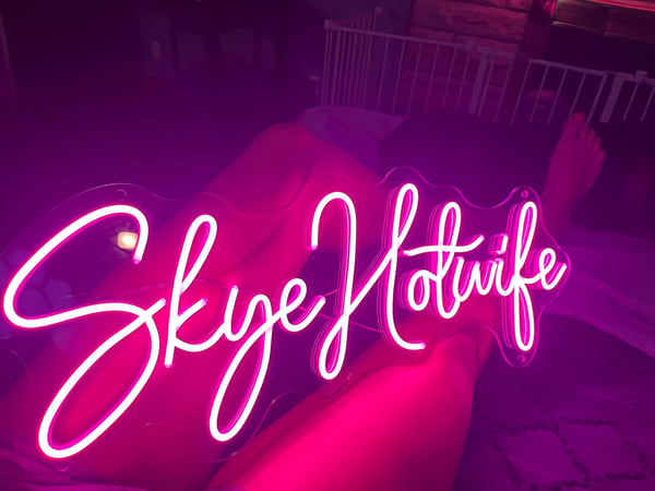 Pink neon sign displaying 'Skye Hotwife' in a dimly lit room with legs visible in the foreground.