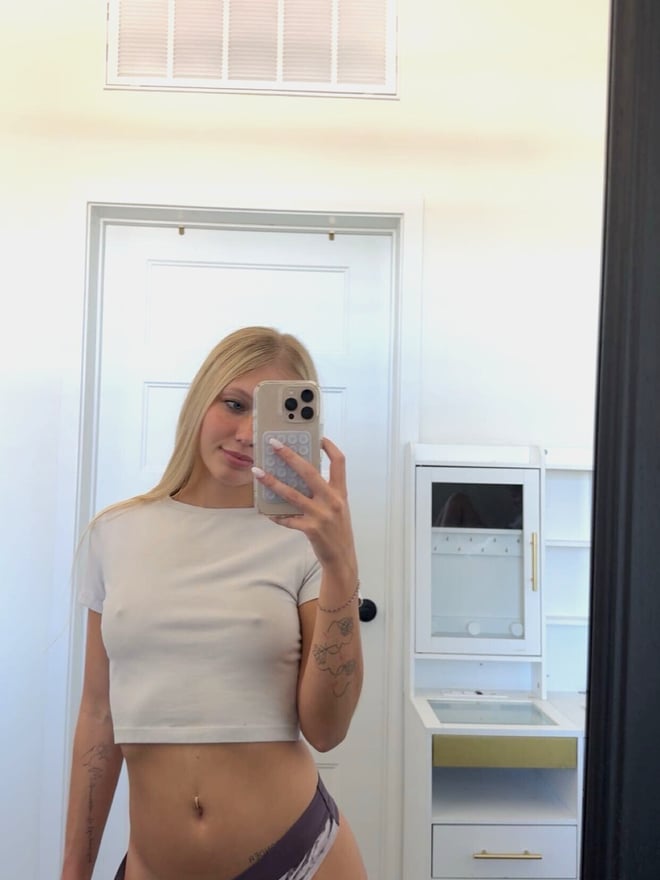 A blonde woman takes a mirror selfie indoors while wearing a light-colored crop top and patterned bottoms.