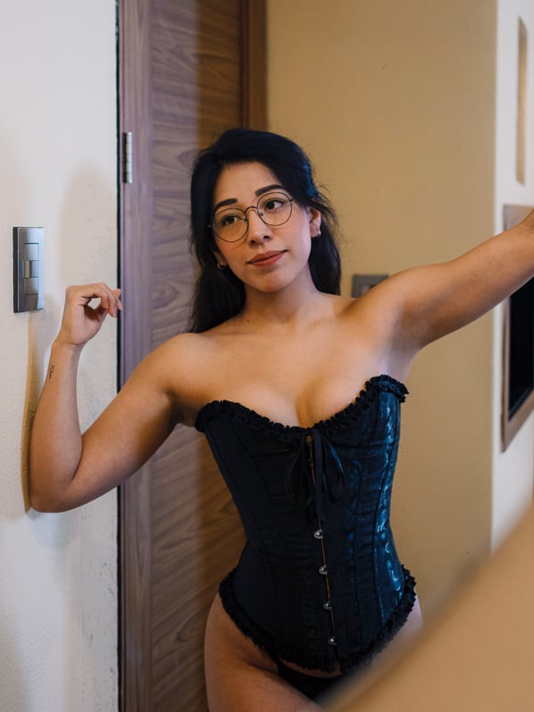 A woman with glasses and long dark hair flexing her arm in a black corset near a wooden door.