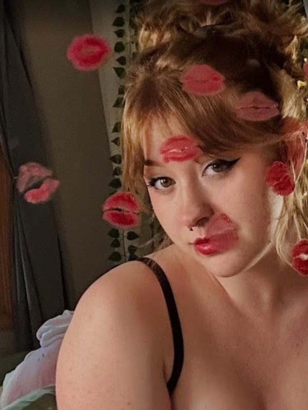A person with voluminous blonde hair and floating pink kiss graphics, posing playfully near a window with greenery in a softly lit room.
