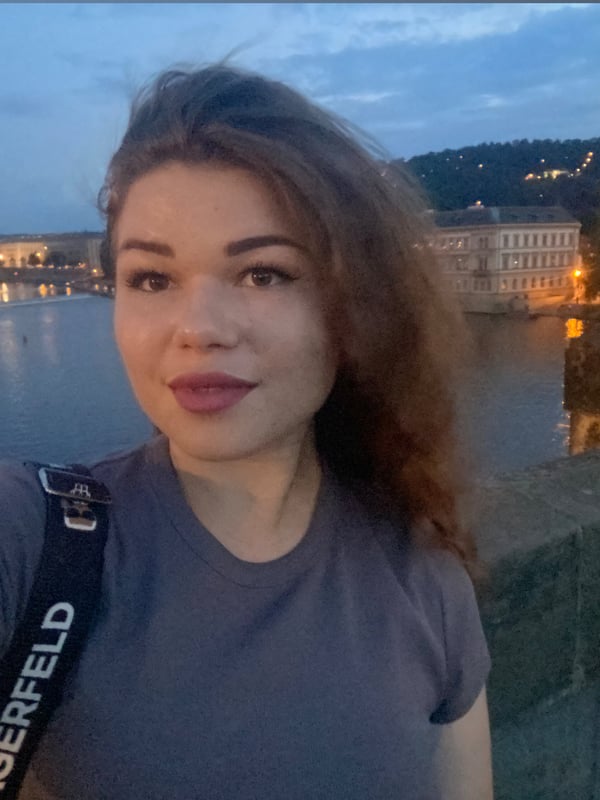 Young woman with brunette hair smiling for a selfie on a bridge at dusk with a river and historic buildings in the background.