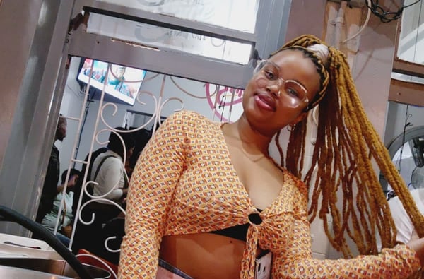 A woman with long braided hair wearing a patterned open blouse and glasses, posing confidently indoors near a window with decorative bars.