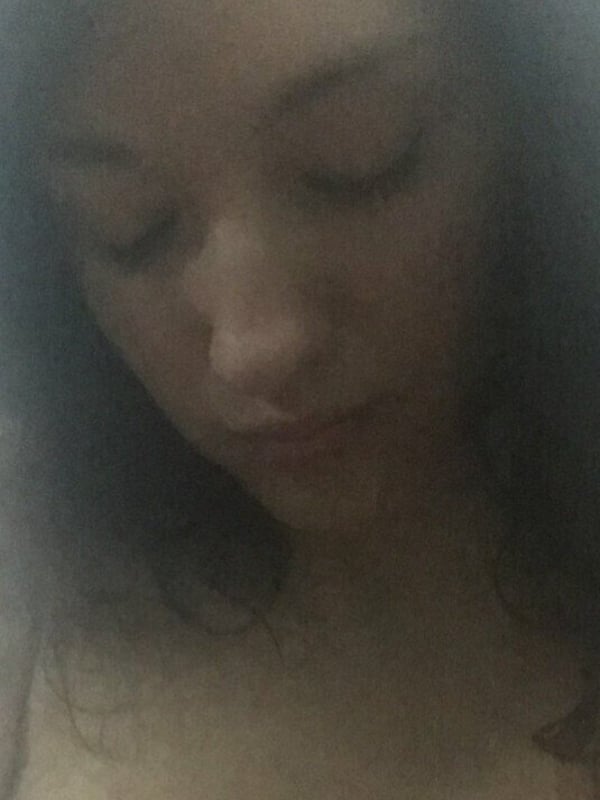 A young woman with long dark hair looking down thoughtfully in a low-light indoor environment.