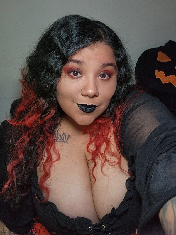 A woman with wavy dark red hair and bold makeup posing with a smiling expression, wearing a black lace top, next to a pumpkin decoration.