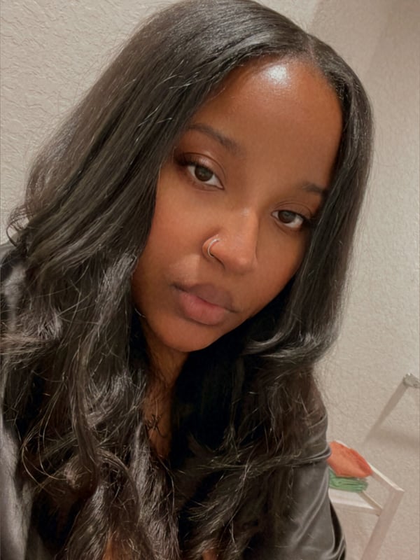 Young woman with long dark hair posing for a close-up selfie indoors with a neutral wall behind her.