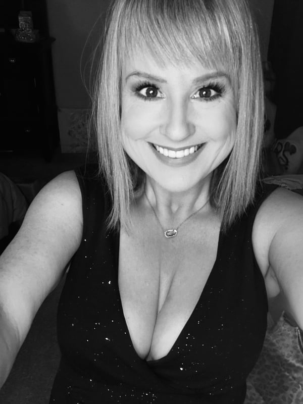 Smiling woman with blonde hair and bangs taking a black-and-white selfie indoors, wearing a low-cut black top.