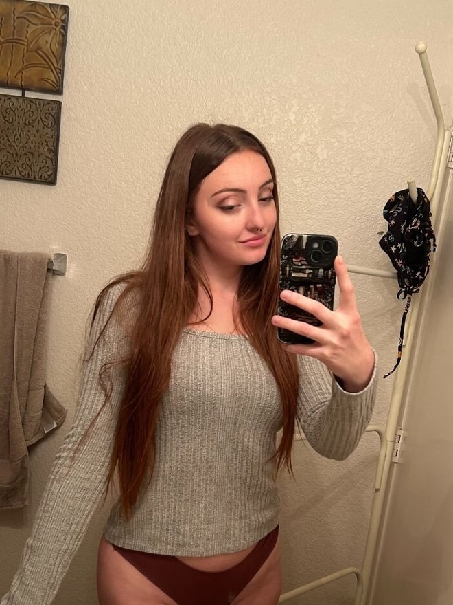 A woman with long reddish-brown hair taking a mirror selfie while wearing a casual knit top.