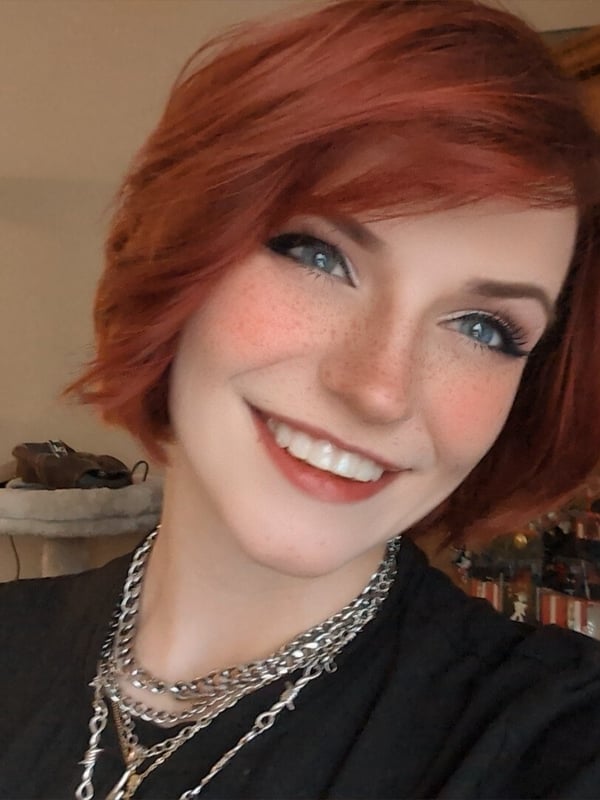 Close-up portrait of a smiling woman with short red hair, blue eyes, freckles, and layered necklaces, posing indoors with wooden furniture in the background.