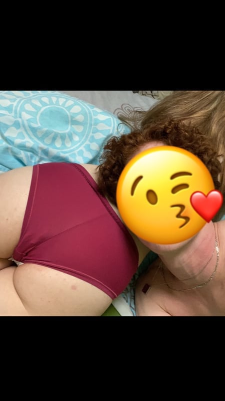 A woman with curly hair lying on a bed, wearing maroon underwear and posing playfully with emojis.