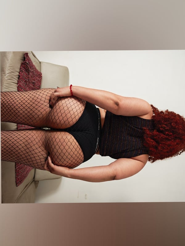 A woman with curly red hair stands facing away, posing confidently in fishnet stockings and dark shorts near a couch with a red blanket.