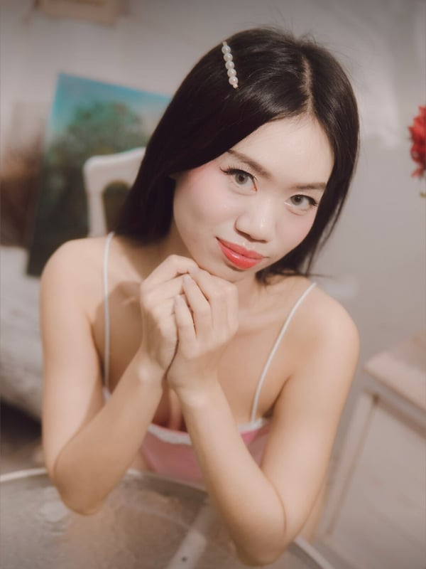 A young woman with long dark hair and a hair clip, smiling softly while sitting with hands clasped, wearing a light-colored top in a cozy room.