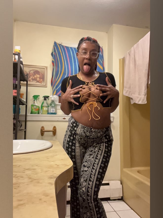 A woman with braided hair making a funny face, standing in a bathroom wearing a black top and patterned pants.