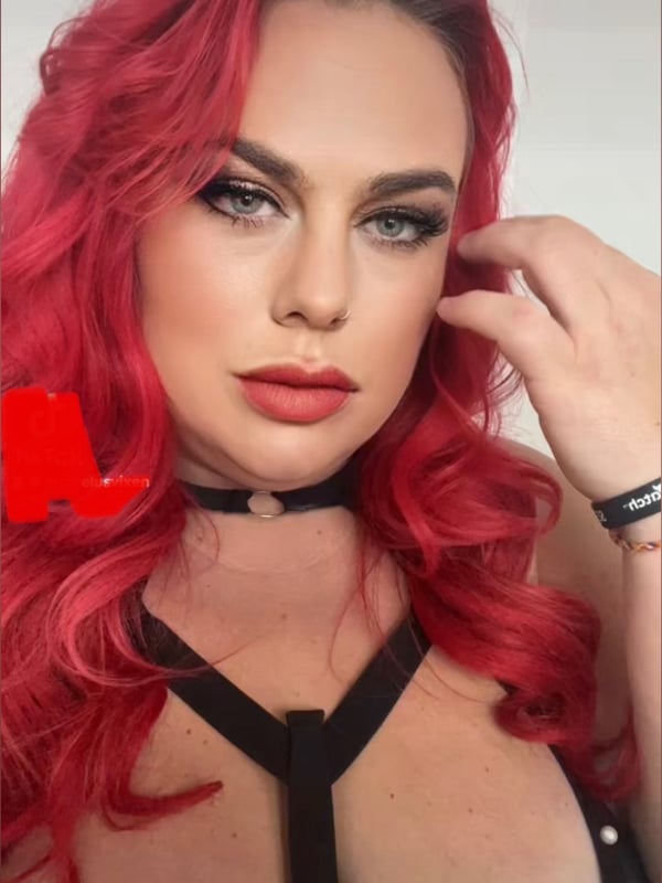 Close-up portrait of a woman with vibrant red wavy hair, bold makeup, and a choker necklace, smiling confidently against a plain background.