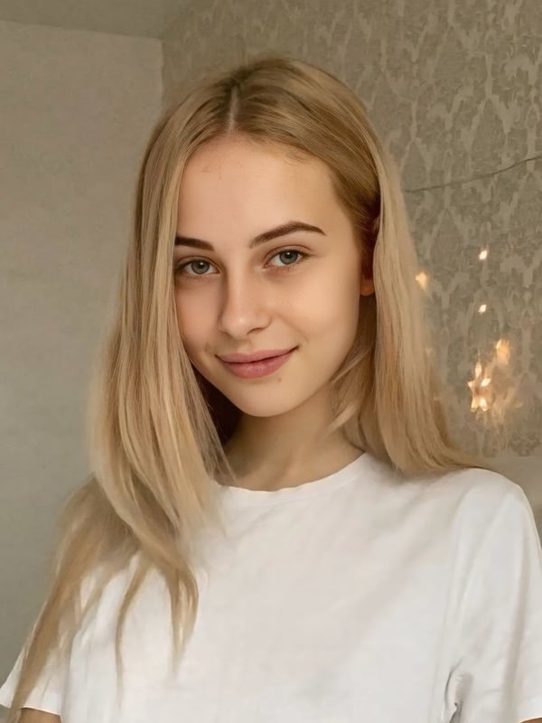 Close-up portrait of a young woman with long blonde hair smiling softly against a textured wall with string lights in the background.