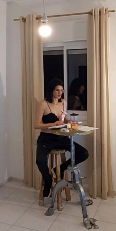 A woman with dark hair sits at a small table by a window at night, writing in a notebook with a lamp illuminating the scene and beige curtains in the background.
