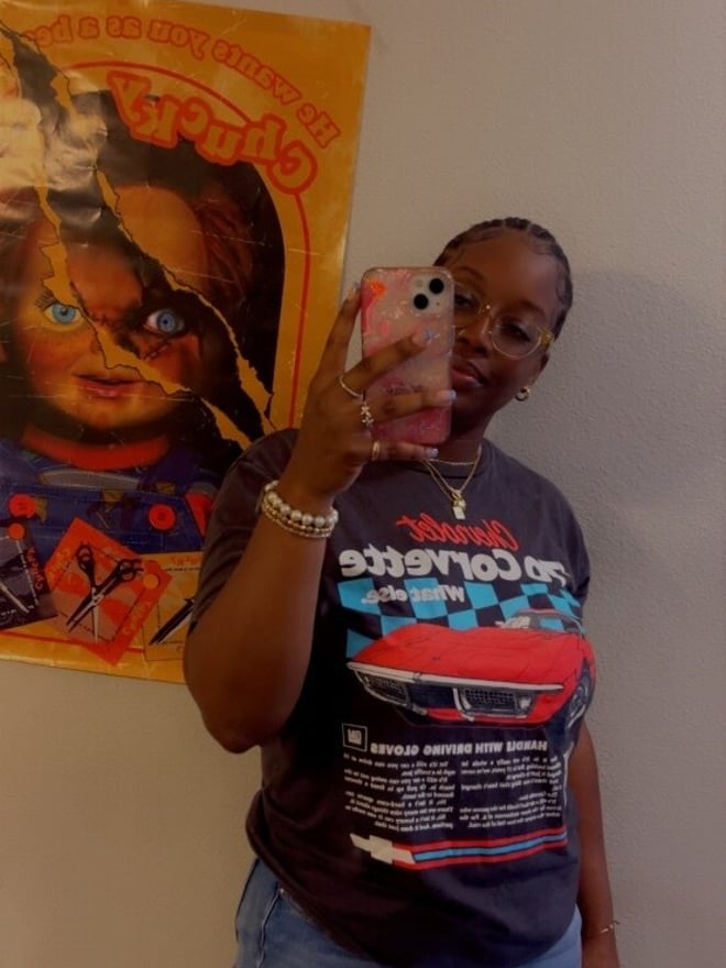 A woman with braided hair smiling while holding her phone, standing in front of a Chucky movie poster and wearing a graphic t-shirt with jeans.