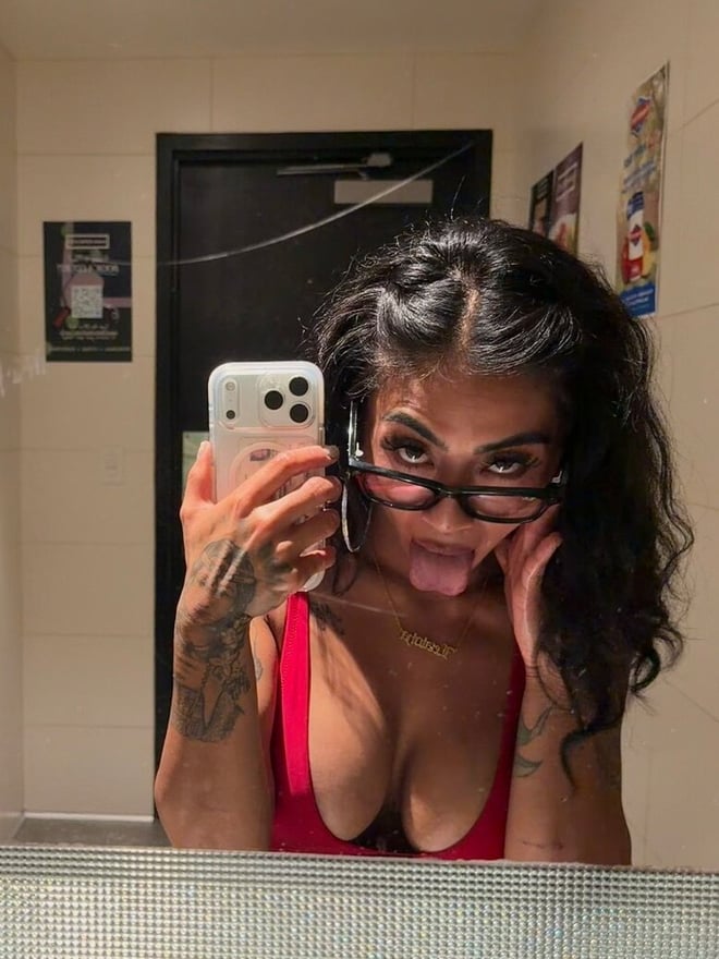 A woman with dark hair and glasses takes a mirror selfie in a bright pink top.