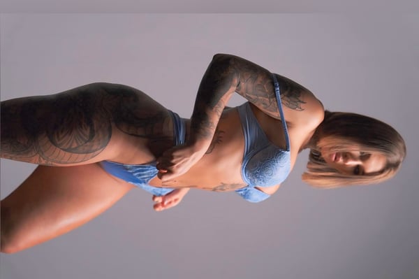 A woman posing confidently in blue lingerie against a gray background, showcasing her tattoos on her arms and legs.