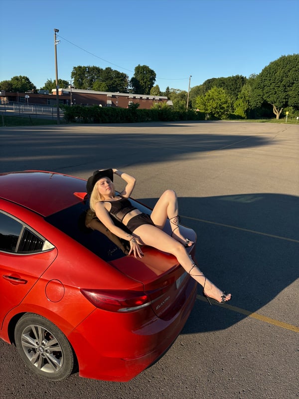 Young woman in black lingerie and hat posing playfully on the roof of a red car in an outdoor parking lot with trees and buildings in the background.