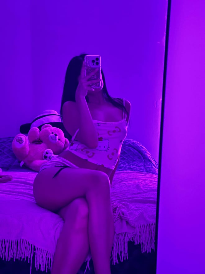 Young woman with long dark hair lying on a pink fluffy rug in a bedroom with pink lighting, holding a phone for a mirror selfie.