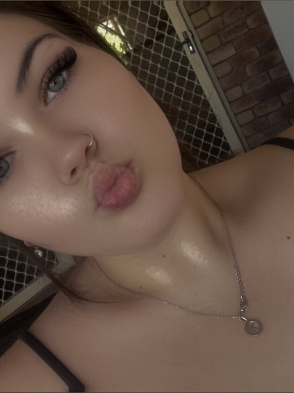 Close-up selfie of a young woman with long dark hair, making a kiss face, wearing a black top against a brick wall background.