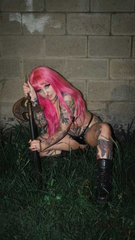 A woman with vibrant pink hair poses confidently outdoors against a graffiti-covered brick wall, holding a staff and wearing knee-high boots.