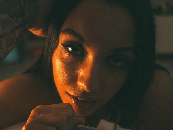 A young woman with tattoos poses dramatically in warm lighting, highlighting her facial features and long dark hair.
