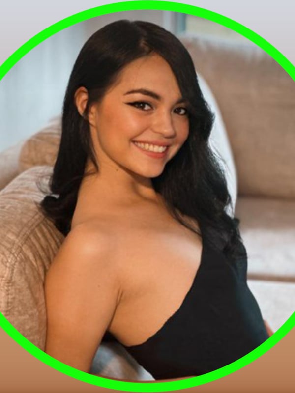 Close-up of a smiling young woman with long dark hair and striking makeup, leaning against a couch in a black top, framed in a green circle.