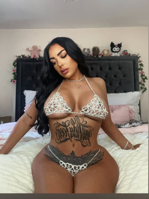 A woman with long dark hair sitting on a bed in a bedroom decorated with plush toys, wearing a white lacy top and jewelry, with tattoos on her body.