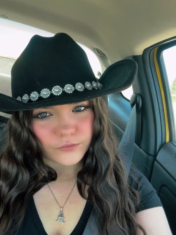 A young woman wearing a decorated black cowboy hat and casual top, sitting in a car.