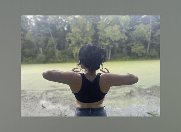 A woman stands with arms outstretched in a black tank top and jeans, facing a green swampy area surrounded by trees under an overcast sky.