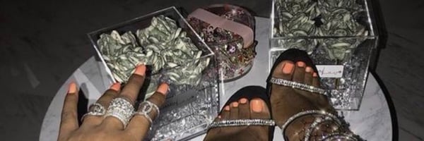 Close-up of manicured hands and high-heeled feet next to boxes of cash and gifts on a table.