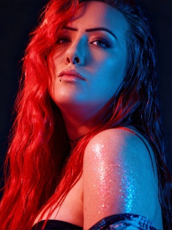 Close-up portrait of a woman with long wavy red hair, dramatic makeup, and blue lighting accentuating her features.