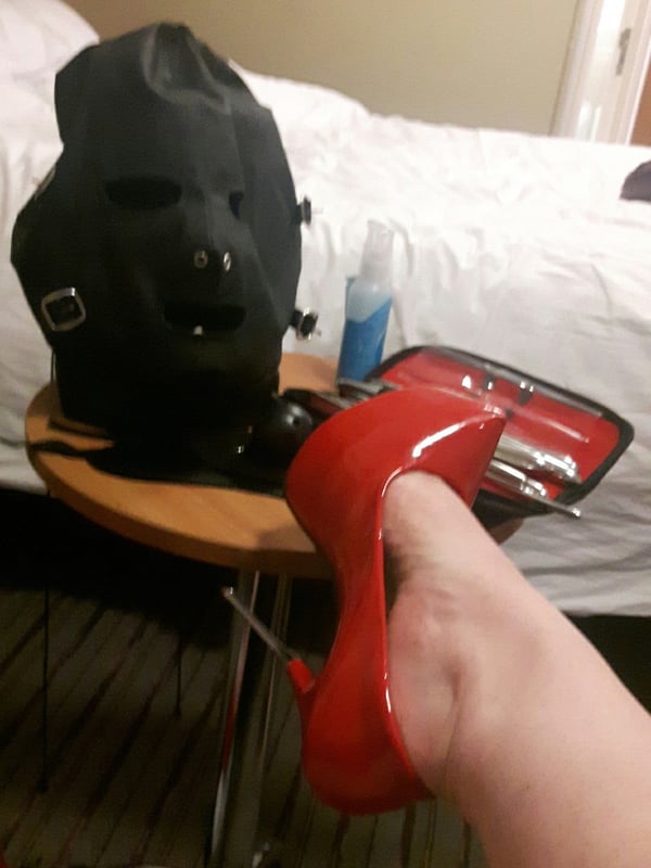 A red high-heeled shoe resting on a table beside a black mask and a red case in a bedroom setting.