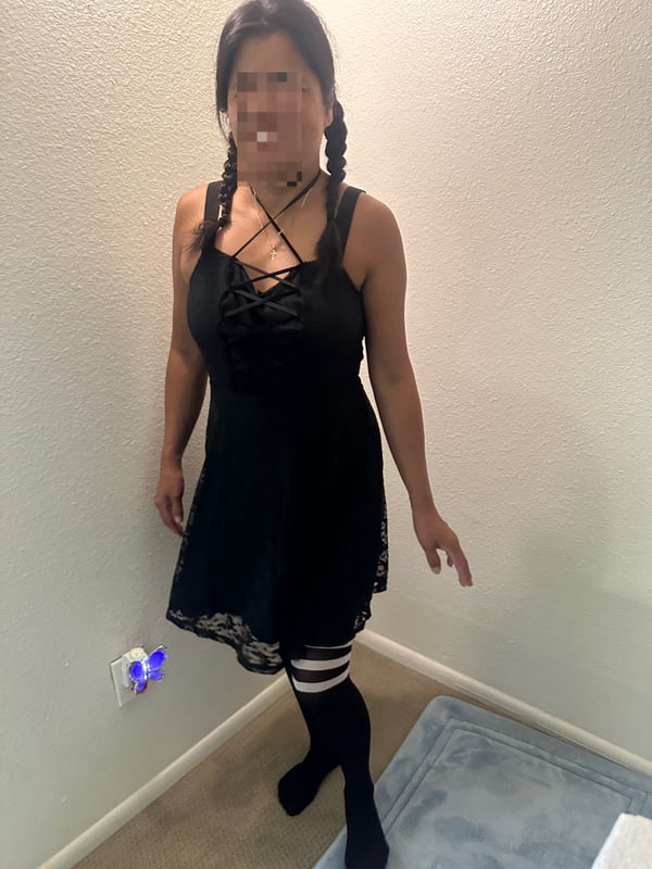 A woman with braided hair and a face blur poses in a black lace top and skirt with striped stockings inside a home.
