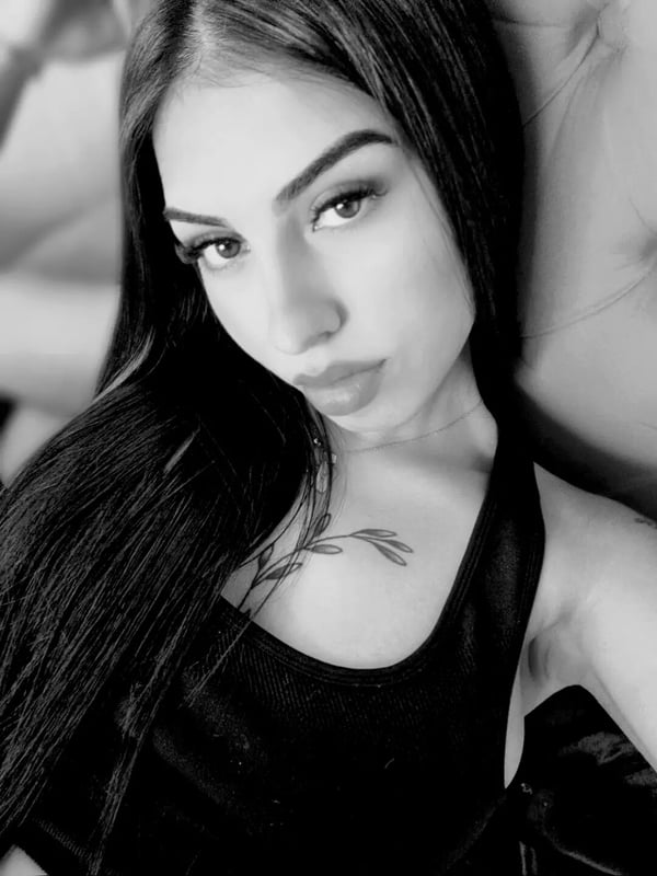 Close-up portrait of a woman with long dark hair, striking makeup, and a subtle tattoo on her chest, leaning against a light background.