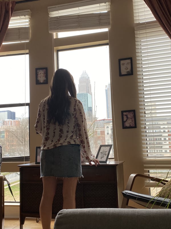 A woman standing by a window in a cozy room with city skyline view, dressed in a skirt and top, looking out.