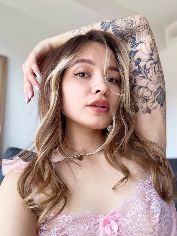 Close-up portrait of a young woman with wavy brown hair and intricate arm tattoos, wearing a delicate necklace and pink top, smiling softly.