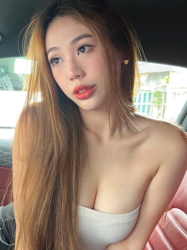 A young woman sitting in a car, smiling at the camera with long dark hair and wearing a white tube top.