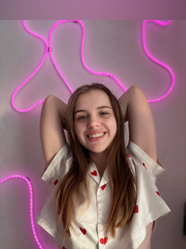 Young woman with long brown hair smiling and posing with arms raised against a neon pink light installation.