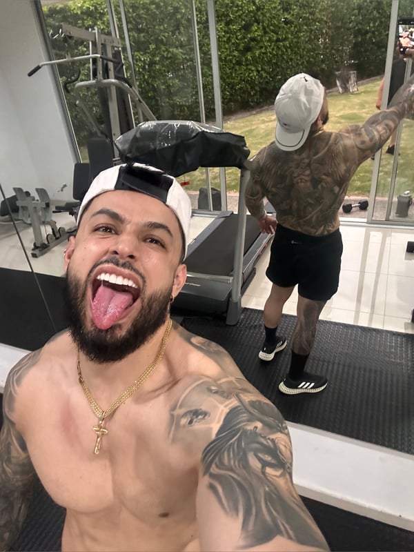 A muscular man with tattoos posing shirtless in a modern gym next to a treadmill, wearing shorts and a cap, with a garden view through glass walls.