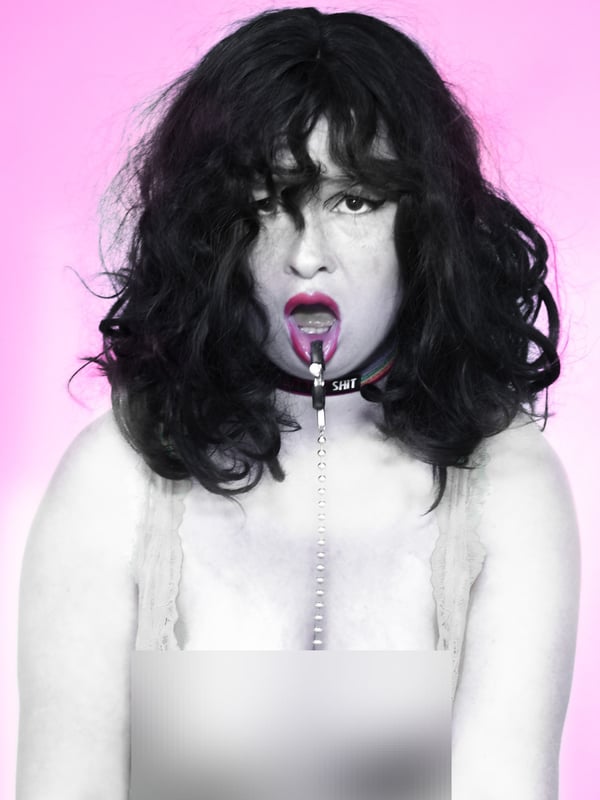 A woman with dark wavy hair poses against a pink background in a black and white image, wearing a choker necklace and expressive makeup.