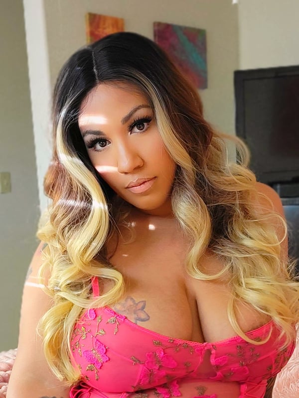 A woman with long wavy highlighted hair wearing a pink lace top, sitting indoors near colorful wall art and furniture.