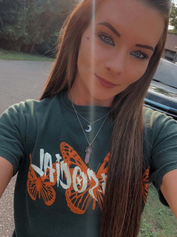 A young woman with long brown hair smiling at the camera outdoors, wearing a green t-shirt with butterfly designs near trees and a driveway.