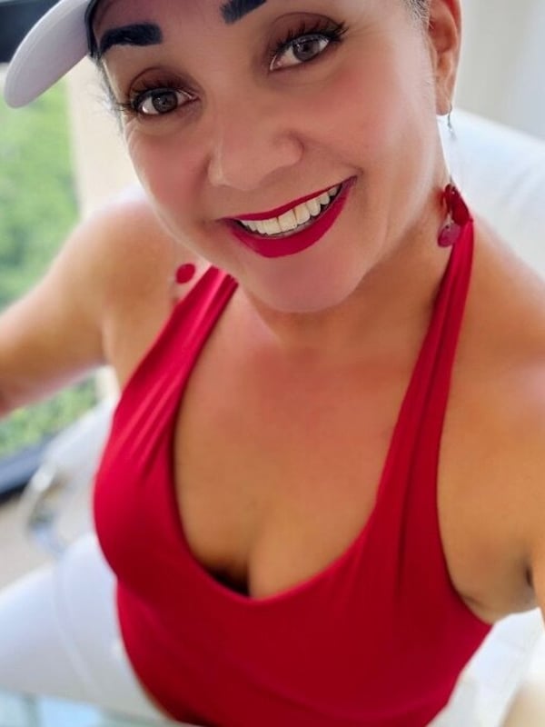 A smiling woman wearing a white cap and red top, posing cheerfully indoors with a window view in the background.