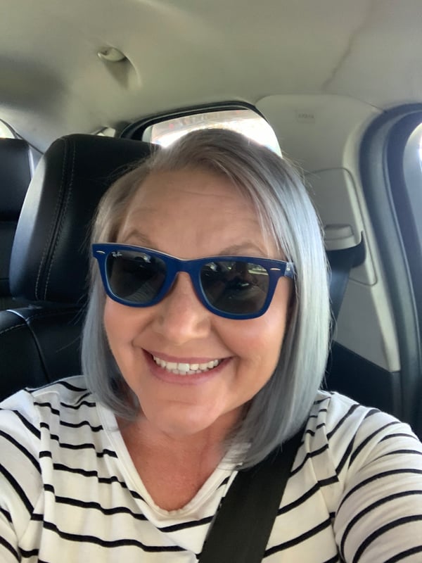 A smiling woman with short gray hair and blue sunglasses sits in a car, wearing a striped shirt and seatbelt.