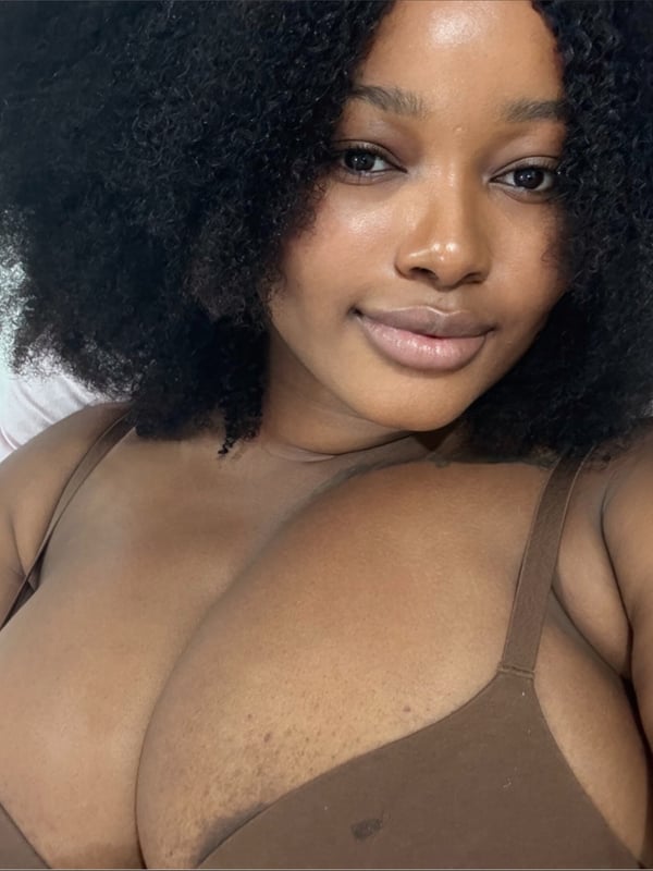 A young woman with curly hair smiling at the camera, wearing a brown top, lying on a bed.