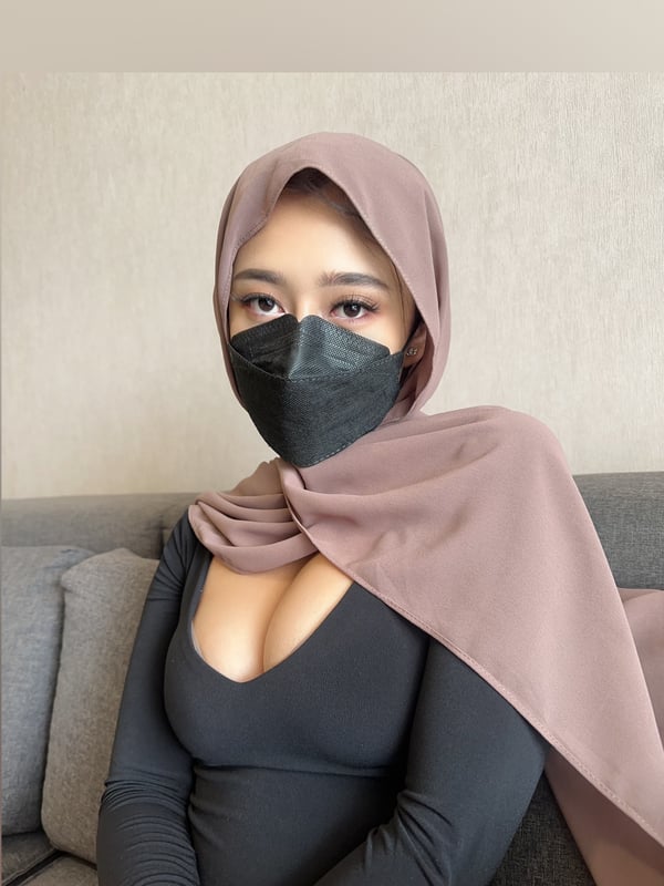 A person wearing a light brown head covering and a dark face mask, seated on a gray couch against a plain wall.