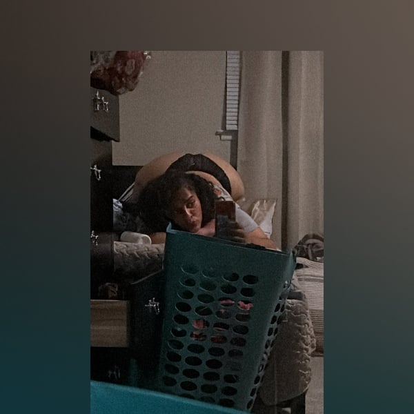 A person lying on a bed in a bedroom with casual clothing and household items nearby.