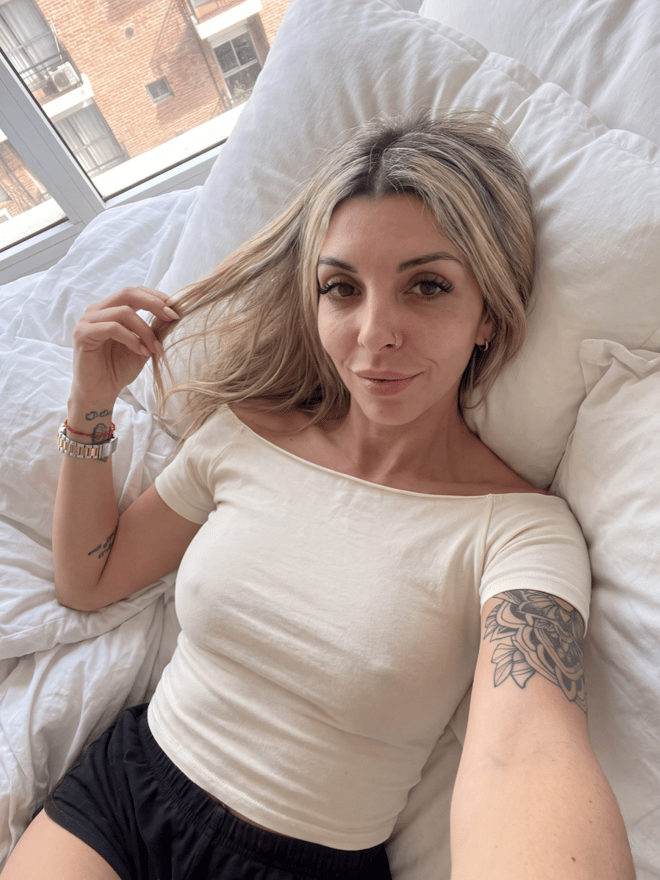 A blonde woman reclining on a bed in casual clothing, with visible arm tattoos and a city view in the background.